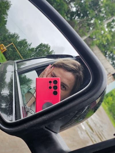 A person is taking a selfie in the side-view mirror of a car. The person's face is partially obscured by their hand, and they are holding a bright pink smartphone. The car appears to be parked on a road in a rural or semi-rural area, with trees and a building visible in the background. The sky is overcast. The image is taken from a slightly low angle, looking up at the mirror. The location is Elovo, Russia.