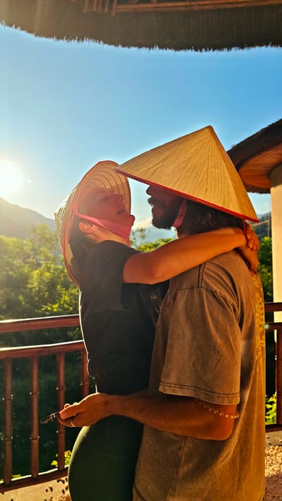 This outdoor portrait captures a joyful and intimate moment between a couple embracing on what appears to be a balcony or terrace in Mu Cang Chai, Vietnam. Both individuals are wearing traditional Vietnamese conical hats (nón lá); the man's hat has a distinct red rim, and the woman's is secured with a pink chin strap.
The woman, positioned on the left, is dressed in a black top and dark green leggings. She looks upwards at the man with a wide, happy expression, suggesting laughter or a broad smile. Her left hand holds a small, dark object, possibly a mobile phone or vape pen. The man, on the right, has a beard and long hair, wearing a light brown or grey t-shirt with a faint pattern on the back. He has a beaded bracelet on his right arm and a ring on his left ring finger. His left arm is wrapped affectionately around the woman's back.
The scene is bathed in warm, golden sunlight from the left, indicative of the golden hour, either late afternoon or early morning. This strong backlighting highlights the subjects and creates a vibrant atmosphere against a clear, bright blue sky. In the background, lush green mountains and trees provide a natural, scenic backdrop typical of Mu Cang Chai. A sturdy wooden railing runs across the foreground and mid-ground. Above and to the right, a structure with a thatched roof is partially visible, suggesting a traditional building or resort. The image conveys strong emotions of affection, happiness, and connection between the couple. No discernible text is visible within the frame.