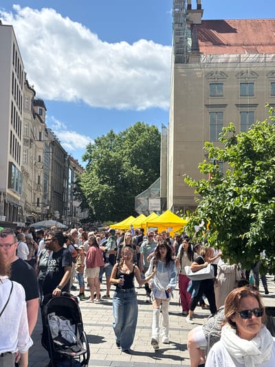 In Munich, Germany, a street fair is underway on a sunny day with white clouds in the sky. The scene is bustling with a crowd of people walking along a paved street lined with buildings. Yellow tents are set up, possibly for vendors or exhibits, and green trees provide shade. Some buildings are under construction, indicated by scaffolding. The focus is on the diverse group of people, varying in age and attire, enjoying the outdoor event.