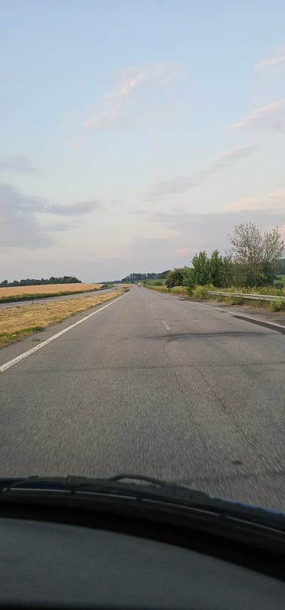 This is a forward-facing, eye-level shot of a road from the perspective of a vehicle in Ulianivka, Ukraine. The road is asphalt and has a white line on the left edge and dashed white lines in the center. On the left side of the road, there is a dry, golden field of crops. On the right side, there are green trees and vegetation, as well as a metal guardrail. In the distance, there are more trees and rolling hills under a pale blue sky with scattered clouds. A single red-and-white truck is visible in the far distance on the road. The foreground shows the edge of a car's dashboard and windshield wipers.