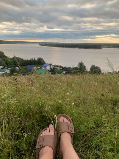 The image depicts a tranquil scene with a person's feet in brown slides resting on a grassy slope overlooking a wide river. In the distance, a village with colorful houses, including ones with blue and green roofs, is nestled along the riverbank. The sky is overcast with dramatic clouds, but rays of sunlight break through, illuminating the water and the horizon. The foreground is dominated by tall, dry grass and scattered wildflowers, suggesting a rural or natural setting, possibly on the outskirts of the village of Olkhovka, Russia. The overall atmosphere is one of peaceful contemplation and natural beauty.