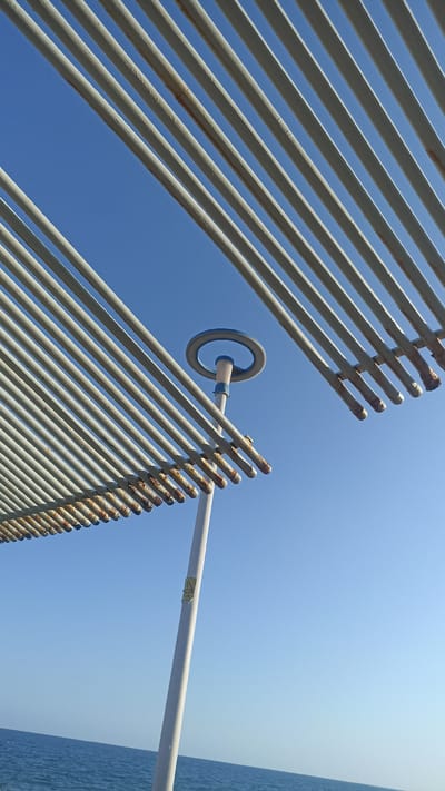 This is a low-angle, outdoor, vertical, eye-level shot of a modern outdoor structure with a clear blue sky and a body of water in the background. The structure appears to be part of a beachside or seaside amenity, possibly a covered walkway or seating area, given the presence of the ocean.
The primary subjects are the parallel white metal beams of the structure, which create a striped pattern against the sky. These beams are angled from the bottom left and top right of the frame, converging towards the center. Some of the beams show signs of rust on their ends, suggesting exposure to the elements.
In the upper central part of the image, a white pole rises, supporting a circular element with a blue outline. This could be a decorative feature, part of a lighting fixture, or a signage holder. A yellow triangular sticker with black text is visible on the pole, but the text is too small to be legible.
The setting is clearly outdoors, with a vast expanse of clear blue sky occupying most of the frame. Below the structure, a calm, deep blue ocean stretches across the bottom of the image. The lighting suggests it is daytime, likely midday or afternoon, with bright sunshine. The weather appears to be clear and sunny.
There are no people visible in the image, and no specific activity is taking place. The overall mood is one of quiet and perhaps a slight sense of neglect due to the visible rust, contrasting with the otherwise clean and modern design of the structure and the serene natural backdrop. The location cues, such as the ocean and the architectural style, are consistent with a seaside resort or public area.