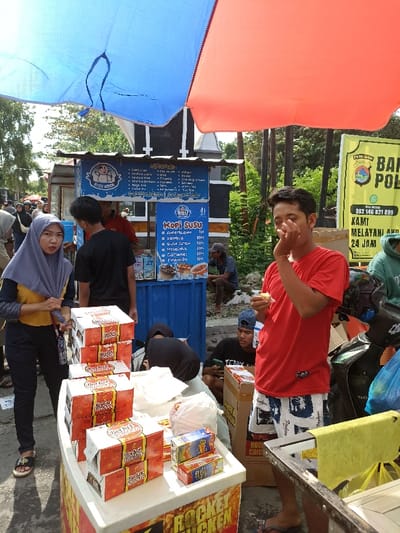 This image depicts a lively outdoor market scene in Mataram, Indonesia, under bright daylight. Two large parasols, one blue and one red, partially frame the top of the image, providing shade over the area.
In the foreground, a white vendor cart showcases stacks of red-and-white boxes labeled "ROCKET CHICKEN." A woman in a grey hijab and yellow long-sleeved top stands on the left, looking towards the camera. On the right, a young man in a red t-shirt and patterned shorts is holding food to his mouth, possibly squinting from the sun.
Behind the foreground cart, a blue food stall is visible, prominently displaying menus for "KOPI SUSU" (Coffee Milk). The menu lists various coffee and milk drink options, such as Americano (7K), Vanilla (10K), Caramel (10K), and Tiramisu (10K), along with other items from a stall potentially named "BUBU MOOK." A man in a black t-shirt stands behind this blue stall. Further back, more people are gathered, some seated, contributing to the bustling atmosphere of a street food area.
In the background, a yellow banner with a police logo is displayed, bearing text that reads "BAP POL," a phone number "082 146 821 899," and the phrase "KAMI MELAYANI AND 24 JAM" (We serve you 24 hours). A motorcycle is also visible on the right side of the frame. The overall scene suggests a typical, busy day at an Indonesian street market, with vendors selling food and drinks to customers.