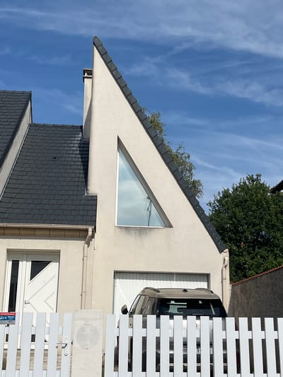 The image displays a modern house with a distinctive triangular window, situated in Chevilly-Larue, France. The house features a dark tiled roof and a beige stucco exterior. A white picket fence runs across the foreground, with a portion of a silver SUV visible behind it. The sky is blue with scattered white clouds. In the distance, lush green trees are visible. A residential street environment is suggested by the presence of houses and fencing. The lighting indicates it is daytime.