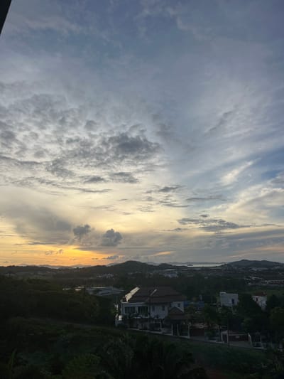 This is an eye-level, wide shot photograph taken from an elevated viewpoint, overlooking a town and surrounding landscape. The sky is filled with a dramatic mix of clouds, with the sun setting or rising on the horizon, casting a warm yellow and orange glow. The clouds are varied in texture and color, ranging from dark gray to wispy white, with some areas illuminated by the setting/rising sun.
Below the sky, a landscape unfolds, featuring rolling hills covered in dense green vegetation. In the mid-ground, houses and buildings are visible, some with red-tiled roofs and white walls, suggesting a residential or developing area. A body of water, possibly the sea or a large bay, can be seen in the distance, with hills forming a backdrop. The overall impression is a tropical or sub-tropical environment.
The specific location is in the city of Ratsada, Thailand. The image captures a view of the urban development interspersed with natural greenery, under a striking sky. There are no people or animals visible in the photograph. The focus is on the landscape and atmospheric conditions during twilight.