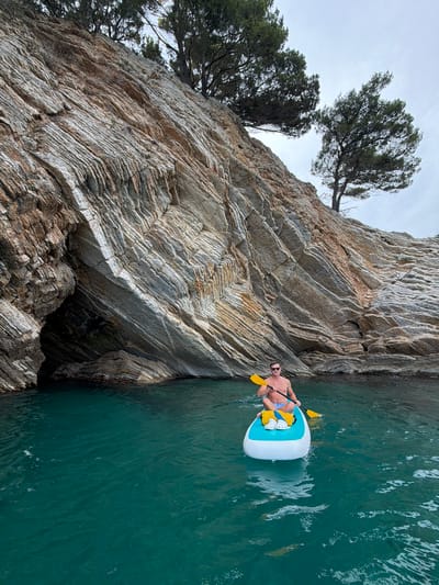 A shirtless man wearing blue and white patterned swim trunks and sunglasses is paddleboarding in the turquoise waters of Petrovac centar, Montenegro. He is holding a yellow paddle. The paddleboard is white with a light blue accent on the top and has two yellow flotation devices strapped to it. The man is positioned on the right side of the frame, while a steep, rocky cliff face dominates the left side. Trees are growing out of the top of the cliff. The sky is overcast. The overall impression is one of a leisurely day spent enjoying the natural beauty of the Montenegrin coast.