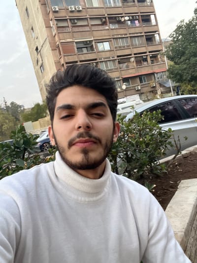 A young man with dark hair, dark eyes, a short beard, and a mustache is depicted in a close-up selfie, wearing a light-cream turtleneck sweater and looking directly at the camera with a neutral expression. The scene is located outdoors in Al-Qanawat Municipality, Syria. Behind the man, green bushes and a section of a light-colored stone barrier are visible. A silver sedan is partially obscured by the bushes on the right, with another darker vehicle visible further back on the left. The prominent background element is a multi-story, aged, concrete-colored building featuring numerous windows, balconies, external air conditioning units, and satellite dishes; laundry is visible hanging on some balconies. To the far left, a narrower, taller building is partially visible. The sky is uniformly overcast and grey.