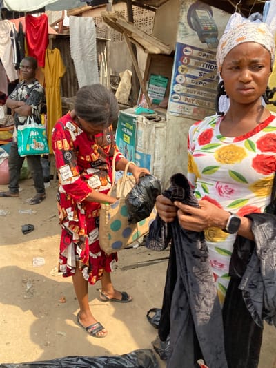 This image depicts an outdoor market scene in Ura Nta Umuarandu, Nigeria, under bright daylight.
Three individuals are visible. In the foreground right, an adult female with dark braids and a white patterned headwrap wears a white, long-sleeved shirt with a red and yellow floral pattern. She holds multiple pieces of black fabric, possibly velvet, in her hands, appearing to inspect them. A dark digital watch is present on her left wrist. Her gaze is directed towards the left side of the frame.
In the mid-ground, slightly to the left, another adult female is bent over, looking into a light brown tote bag with blue polka dots. She wears a red, long-sleeved dress featuring a repeating pattern of "GUCCI Bloom" text and what appear to be perfume bottles. A black plastic bag is held in her right hand above the tote bag. She is wearing dark flat sandals.
In the background left, a young male or teenager stands with crossed arms, looking towards the camera. He wears a short-sleeved patterned shirt and dark trousers, with dark sandals. He holds a light green tote bag with an illustrated design in his left hand.
The environment consists of unpaved, sandy ground with scattered debris, including small pieces of paper and black plastic bags. Makeshift market stalls are visible in the background, constructed from wood, corrugated metal sheets, and draped fabric. Hanging clothing items, including a red top, a yellow dress, and other garments, are displayed for sale. Prominently displayed signage on the right includes a blue and white poster depicting a POS terminal and listing services such as "OPEN ACCOUNT," "WITHDRAW," "SEND MONEY," "DEPOSIT," "BUY RECHARGE," and "PAY BILLS" (including DStv and GOtv). A smaller green and white sign below it states "ATM Card - ONE Minute." Another partial sign above includes "PAY WITH TRANSFER" and a phone number.