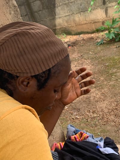 The image shows a person wearing a brown headscarf and a yellow shirt, with their hand covering their face. The background consists of a dirt ground with some scattered leaves and a concrete wall. In the foreground, there is a pile of clothes, including a grey shirt with colorful stripes and black pants. The scene appears to be outdoors, possibly in a residential area. There is no specific indication of the city of Agban, Nigeria within the image itself.