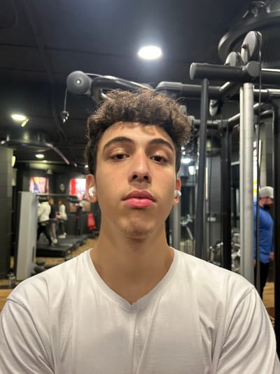 A young man with curly hair and light skin is looking at the camera. He's wearing a white t-shirt and what appear to be wireless earbuds. The image seems to be taken in a gym, with exercise equipment visible in the background. Another person, wearing a blue shirt and a white cap, is also visible, possibly working out. The lighting suggests an indoor setting with artificial lights.
