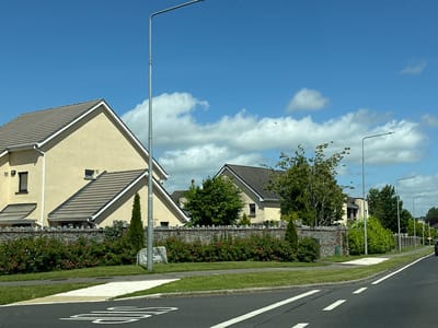 The image depicts a residential street in Castledermot, Ireland, on a clear day with a bright blue sky and scattered white clouds. The foreground shows an asphalt road with white dashed lane markings and a pedestrian crossing. To the left of the road, a well-maintained green lawn is visible, separated from a stone wall by a sidewalk. The stone wall is topped with a row of green bushes and colorful red flowers. Behind the wall, several houses with beige exteriors and brown tiled roofs are visible. Tall streetlights line the right side of the road, interspersed with trees and more green landscaping. A stone marker with "AR LANDS HILL" is placed near the sidewalk. The overall scene suggests a quiet, suburban environment.