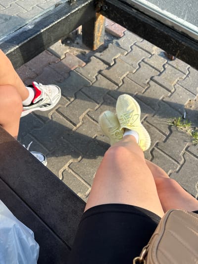 The image shows a close-up of two people's legs and feet, sitting on a textured dark surface. One person is wearing light yellow sneakers with white socks and black shorts. The other person is wearing white and red athletic shoes with white socks. The background is a grey paved surface, suggesting an outdoor setting. Sunlight casts shadows, indicating it is daytime. The image has a casual and relaxed feel. No visible text is present in the image. The location context provided is Mytishchi, Russia.