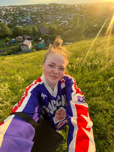 The image is a selfie taken outdoors in the city of Olkhovka, Russia, likely during golden hour due to the strong sunlight and lens flare. The foreground features a young woman with blonde hair, freckles, and a blue, white, and red jersey that appears to be a sports uniform. She is smiling and looking at the camera. Her jersey has Cyrillic text and a coat of arms. A black bag is visible over her shoulder, with a purple strap or cover. The woman is standing on a grassy slope covered in small white flowers and some yellow buttercups. In the background, a panoramic view of a village or town is visible, characterized by numerous small houses with various colored roofs, interspersed with trees and green spaces. The sunlight casts long shadows and illuminates the landscape, creating a warm and hazy atmosphere. The houses appear to be predominantly wooden or made of brick, with some featuring more modern construction. The overall impression is of a rural or semi-rural setting bathed in the warm glow of sunset.
