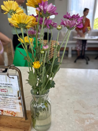 The image shows a glass vase of purple and yellow flowers sitting on a table, likely in a restaurant. A wooden stand with a sign for Amino is visible in the foreground. The sign advertises a bonus program and asks customers to fill out a form with their name. In the background, blurred people can be seen sitting at tables, including a child with an orange shirt and another table with a person sitting at it. The scene appears to be indoors, with natural light coming in from a window or opening, and is likely located in Ungasan, Indonesia due to the sign referencing the Amino app, which is common in the region.