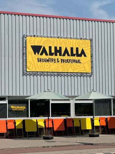 This is a photograph taken outdoors, likely during the day given the bright sunlight casting shadows. The scene features the exterior of a modern building with a grey, corrugated metal facade. Attached to this facade is a large, bright yellow banner with bold black lettering. The text on the banner reads "VALHALLA" in large, stylized font, followed by "BROUWERIJ & PROEFILOKAAL" in a smaller, more conventional font. This indicates that Valhalla is a brewery and tasting room.
To the right of the banner, the building has a series of large windows with dark frames. Outside these windows, there is a row of colorful outdoor seating. The furniture consists of tables and chairs, primarily in bright orange and yellow. Some of the chairs and tables are visible through the windows as well. Partially obscuring some of the windows and the upper portion of the banner are light green, angled awnings or umbrellas, suggesting an outdoor seating area that can be shaded.
The sidewalk or pathway in front of the building is paved with grey and brown tiles, and there's a dark strip of material running along the edge of the pathway. The sky visible in the upper left corner is clear and blue, with no clouds. The overall impression is that of a commercial establishment, possibly a bar or restaurant, with an inviting outdoor seating area. The setting appears to be urban, given the architectural style of the building and the presence of a paved walkway. No people are visible in the image.