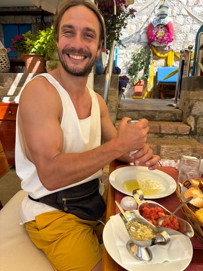 A smiling man with blonde hair and a beard sits at a table with food and drinks. He is wearing a white tank top and yellow shorts, with a fanny pack around his waist. In front of him is a white plate with a dish of food, a glass of water with ice, a basket of bread, and two small metal dishes containing chopped tomatoes and a finely chopped mixture of garlic and herbs. The man is holding a small metal container in his hands.
The setting appears to be an outdoor restaurant or cafe, with stone walls and steps in the background. There are potted plants and flowers visible, adding to a pleasant ambiance. A red and white life preserver with flowers hangs on the wall, along with a yellow cloth draped behind it. The overall scene suggests a relaxed meal in a sunny location. This scene is likely in the city of Pržno, Montenegro, based on the architectural style and lush vegetation typical of the area.
