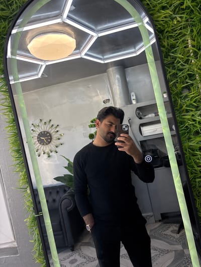 A man takes a selfie in a mirror. The mirror is surrounded by artificial green grass. The environment reflected in the mirror includes a gray wall, a black chair, and a decorative wall clock. The man is wearing a black sweater and holding a phone. The location of the photo in Dolni Tsibar, Bulgaria is not discernible.