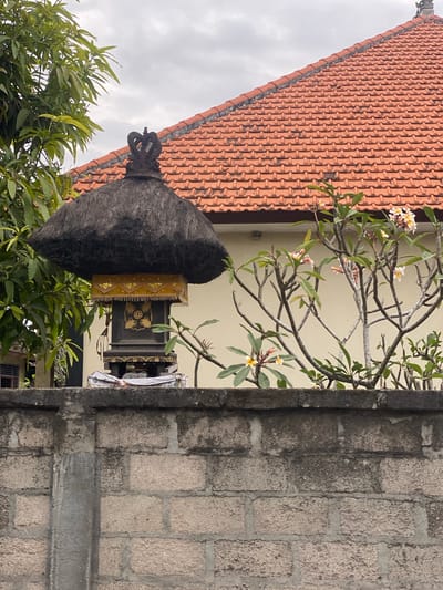 In Nusa Penida, Indonesia, a Balinese offering shrine with a thatched roof and a dark, decorative top stands behind a grey concrete block wall. A frangipani tree with white and pink flowers is in front of a cream-colored building with a red tile roof. The sky is overcast.