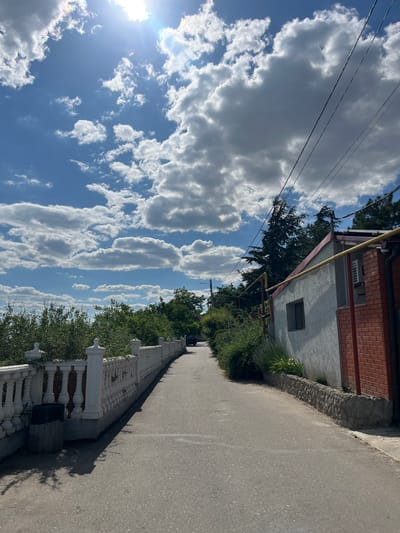 The image depicts a street scene in Kryzhanivka, Ukraine, on a bright, sunny day with scattered white clouds against a blue sky. A paved road curves gently into the distance, bordered on the left by a white ornamental fence with balusters and on the right by a building with a light-colored stucco exterior and a red brick lower section. Trees and green foliage line the left side of the road behind the fence, and more trees are visible further down the road and to the right behind the building. Overhead power lines traverse the upper right portion of the frame. A dark-colored trash receptacle is visible near the white fence on the left.