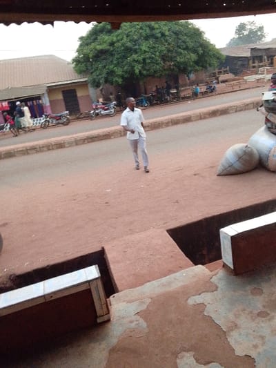 This media file presents a daytime street scene in what appears to be a town or village, likely Ankpa, Nigeria, given the contextual information. The view is slightly elevated, looking down onto a reddish-brown dirt road that dominates the center of the frame.
A man of African descent, dressed in a light-colored, short-sleeved shirt and light trousers, is walking across the unpaved road from right to left. He is the most prominent figure, situated in the middle ground. The road is bordered by a low, concrete curb.
On the left side of the road, a low-rise building with a corrugated iron roof is visible, appearing to be a shop with fabrics or goods displayed outside. Several people, including children, are seen near this building, and a few motorcycles are parked along the side of the road. In the background behind this area, more buildings with similar architecture are present.
To the right of the road, a large, leafy green tree stands prominently, offering shade. Under and around this tree, other people are gathered, and several motorcycles are parked. In the foreground on the right, several large, light-colored sacks (possibly filled with produce or goods) are stacked on the ground next to what looks like a scooter or moped. The immediate foreground also features concrete structures, including a visible open trench or drainage channel.
The overall atmosphere suggests routine daily life in a bustling, semi-urban environment. The lighting indicates it is daytime, possibly with a slightly overcast sky, as shadows are not harsh. There is no discernible text visible in the image.