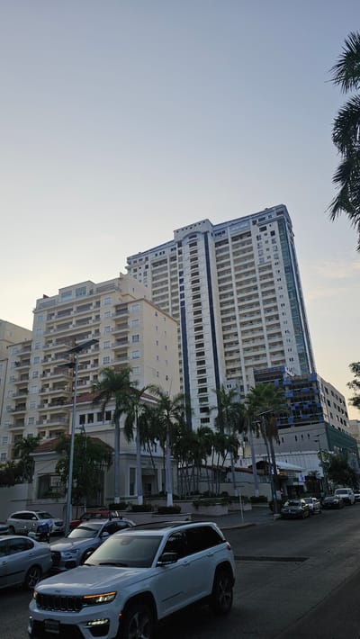 The image shows a street scene in Acapulco, Mexico. Several tall buildings are visible, appearing to be residential or commercial properties with multiple floors and balconies. Palm trees line the street. Parked cars are visible on the right side of the frame. The sky is clear. The scene appears to be taken during daylight hours.