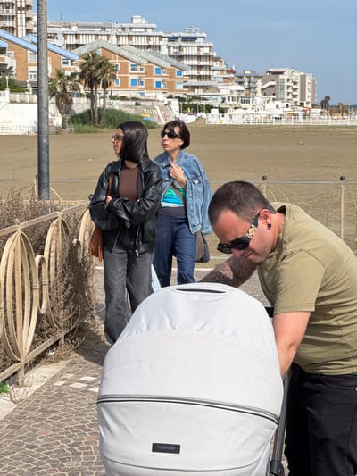 The image shows a group of people on a paved promenade overlooking a beach. In the foreground, a man wearing sunglasses, a light green shirt, and black pants is leaning over a white stroller. Two women are standing behind the man, looking in the same direction. One woman is wearing a denim jacket and jeans, while the other is wearing a black leather jacket, brown top, and dark jeans.

The background features a beach with white sand and a row of buildings in the distance. Palm trees are visible near the buildings.

The location is Nettuno, Italy.