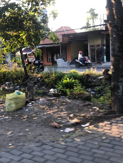 The image depicts a street scene in Kalidawir, Indonesia, characterized by a mix of residential buildings, vegetation, and visible trash. In the foreground, a bright yellow trash bag is prominently placed on the ground next to a waterway. Further into the scene, a person is riding a motorcycle, heading towards the right of the frame. Behind the motorcycle, a small food cart with an umbrella is visible, suggesting a commercial area. The buildings appear to be single-story structures with tiled roofs. The overall environment shows signs of neglect, with debris and vegetation accumulating around the waterway. The lighting suggests it is late afternoon or early morning, with the sun casting long shadows.