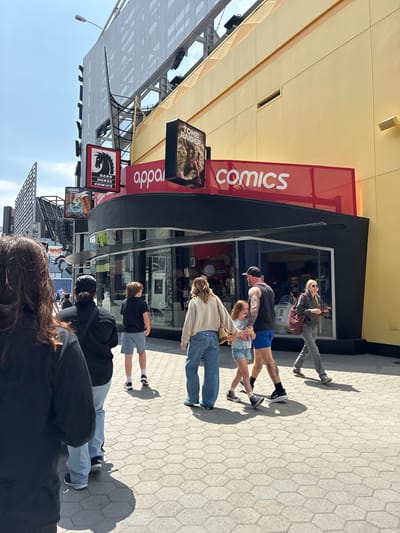 The image depicts an outdoor scene in front of a building with a yellow facade and a red awning that reads "apparel comics." A sign for "DARK HORSE COMICS" with a black horse logo is visible above the awning. Several people are walking on a hexagonal-tiled sidewalk. In the foreground, a person with long brown hair faces away from the camera. Behind them, a group walks towards the building. This group includes a person in a black shirt, a child in a black t-shirt and blue shorts, a man in a black tank top and blue shorts with tattoos, a young girl in a tie-dye shirt and denim shorts, and a woman in a gray hooded jacket and jeans. Further down the sidewalk, another person in a gray jacket and jeans walks away from the camera. The building appears to be a retail establishment, likely a comic book store or a store selling comic-related merchandise, situated in an urban environment. The sky is clear and blue, suggesting fair weather.