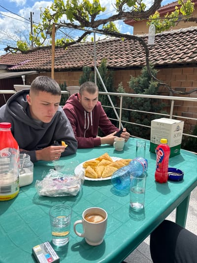 Two young men are sitting at a green plastic table outdoors in what appears to be Graf Ignatievo, Bulgaria. One man is eating something while the other looks at a mobile phone in his hand. There are drinks (including water and juice), food (pastries), and items such as a box of wine and a pack of cigarettes on the table. The background includes a building with a tiled roof and foliage, and the sky is visible.