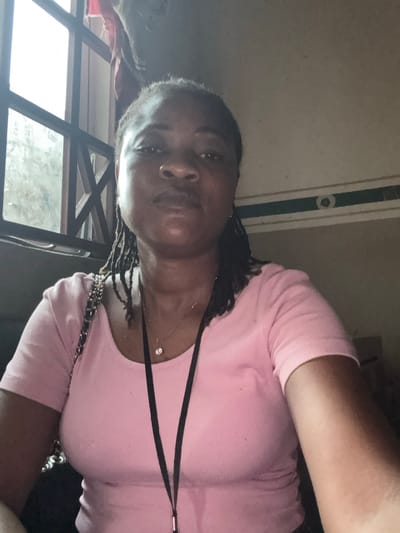 A woman, facing the camera, with dark skin and braided hair is wearing a pink t-shirt. She is wearing a necklace with a diamond pendant. The background appears to be indoors, with a ceiling and a window with a view of foliage and what appears to be a grid-like structure. The lighting suggests it might be daytime. The overall tone of the image is casual.