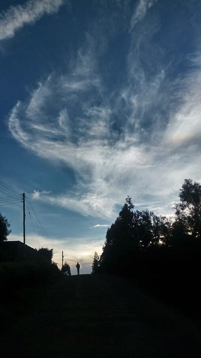 The image shows a scene in Ndeiya, Kenya. The sky is predominantly blue with wispy, cirrus clouds stretching across a large portion of the frame. A power line pole with wires extends upwards from the lower portion of the scene. The horizon is marked by a tree line, silhouetted against a brighter sky, indicating either sunrise or sunset. A single human figure is silhouetted against the light on the horizon. The landscape is otherwise dark, with trees and possibly buildings in silhouette.