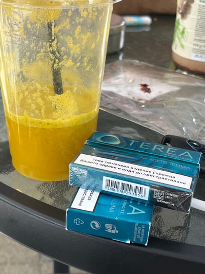 A close-up, eye-level shot captures a clear plastic cup filled with a vibrant yellow, frothy beverage, likely a smoothie or juice, sitting on a dark, reflective, textured surface, possibly a table. A black straw is submerged in the drink. Adjacent to the cup are two packs of Terea Turquoise for IQOS ILUMA tobacco sticks, one stacked on top of the other. The packaging of the tobacco sticks is predominantly turquoise and features branding and warning labels in Bulgarian. The background is blurred but suggests an outdoor setting with a suggestion of a bottle and other objects. No individuals are visible in the frame. The scene is devoid of any discernable Pazardzhik specific landmarks or identifying features.