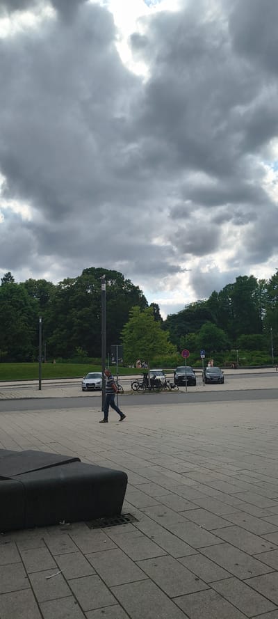 A person walks across a paved plaza with a dark bench in the foreground. In the background, several cars are parked on a street next to a grassy area with trees. The sky is filled with heavy, gray clouds. The scene is located at the Elbphilharmonie in Hamburg, Germany.