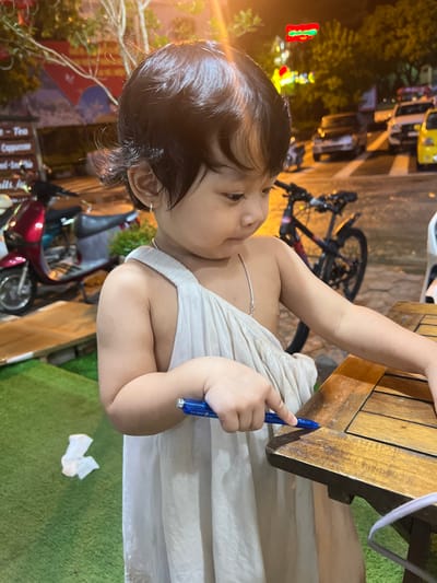 The image shows a young child, appearing to be of Southeast Asian descent, standing next to a wooden table outdoors at night. The child is wearing a simple, off-white garment. They are holding a blue pen and appear to be drawing on the table's surface. In the background, there are out-of-focus elements including a motorcycle, a bicycle, cars, and neon lights, suggesting an urban or commercial setting. The ground in front of the child is covered in artificial green turf, with a crumpled white tissue visible. The location is specified as Bắc Hà, Vietnam.