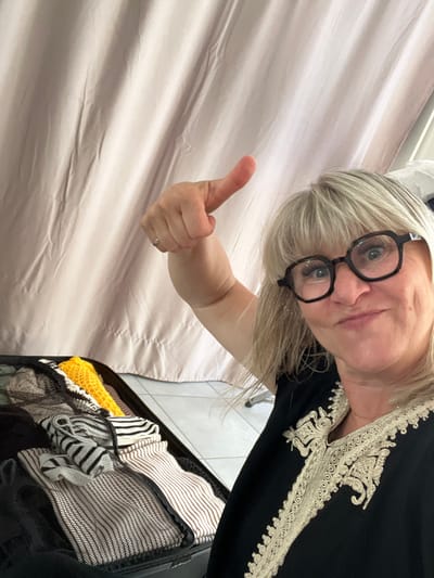In Chambœuf, France, a woman with blonde hair and glasses is taking a selfie. She is wearing a black garment with ornate gold embroidery around the neckline. Her left arm is raised, and she is giving a thumbs-up gesture. In front of her is an open suitcase filled with neatly folded clothing, including items with stripes and a bright yellow knitted piece. A light-colored curtain hangs in the background. The scene suggests preparation for travel.