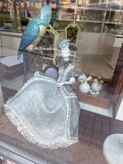 This media file depicts a close-up, angled shot of a porcelain figurine displayed in a shop window. The primary subject is a figurine of a woman in a long, flowing, light-colored gown with intricate ruffles. She is seated and holding a black, ornate mask. Her hair is styled in an elaborate updo with pastel accents.

To the left of the woman, and slightly out of focus, is a blue and green porcelain bird, possibly a parrot, perched on a dark, textured surface. In the background, behind the woman, are several white, spherical ornaments tied with gold ribbon. These ornaments appear to be decorated with small, sculpted figures. The setting is a display window, suggested by the reflections on the glass and the objects visible behind the figurines, which seem to be other shop merchandise and possibly the interior of the establishment.

The lighting appears to be artificial, typical of an indoor display, and no specific time of day or weather conditions are discernible. The overall impression is one of elegance and artistry, showcasing the delicate craftsmanship of the figurines. The location is Karlovy Vary, Czechia, implying a European setting for this shop display. No visible text is present in the image.