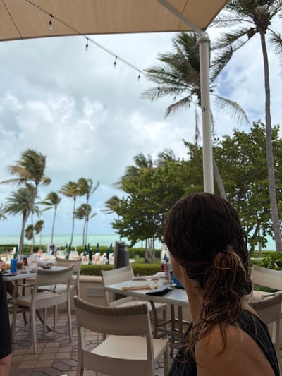 The image shows a view from an outdoor seating area. Palm trees are visible in the background against a cloudy sky. The seating area has white chairs and tables. A person with dark, wet hair is partially visible in the foreground. In the distance, people are gathered near the beach. It is a beachy location on Key Biscayne.