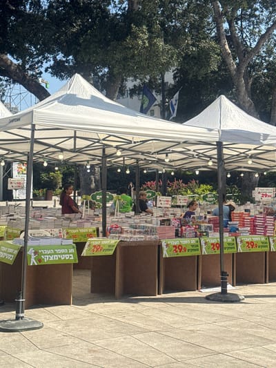 This image shows an outdoor book sale in Ramat Gan, Israel. Several white pop-up tents are set up, displaying stacks of books for sale. The books are neatly arranged on tables and shelves under the tents. Various signs with Hebrew text and prices, such as "29.90", are visible, indicating special offers and the nature of the event.

In the foreground, a portion of a paved walkway and a black tent pole with a stand are visible. Behind the books, there are people browsing and working at the stalls. Some individuals are seen from the shoulders up, engaged in conversations or attending to customers. In the background, lush green trees provide shade, and a glimpse of other structures and flags can be seen. The overall scene depicts a lively book market atmosphere on a sunny day.