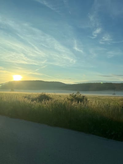 The image shows a landscape at sunrise. In the center of the frame, sunlight breaks through the horizon, behind a ridge of land. To the left of the image, the sky is blue with wispy clouds. To the right of the image, there is a road. The location is possibly in the city of Kastel, Bulgaria, based on provided information.