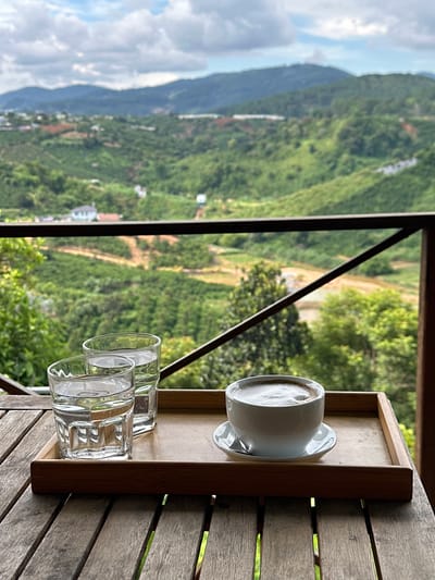 A high-angle medium shot captures a wooden table with a bamboo tray on it, holding two glasses of water and a cup of coffee. The table is made of weathered wooden planks, and the tray is simple and light-colored. The two glasses are clear and filled with water, with light glinting off their surfaces. The coffee cup is white and round, with a frothy beverage inside, and rests on a matching saucer. In the background, a lush green valley unfolds, dotted with houses and fields, leading up to rolling hills and mountains under a cloudy sky. The scene appears to be from a balcony or elevated viewpoint in Thôn 3, Vietnam, offering a panoramic view of the rural landscape. There are no people present in the image.
