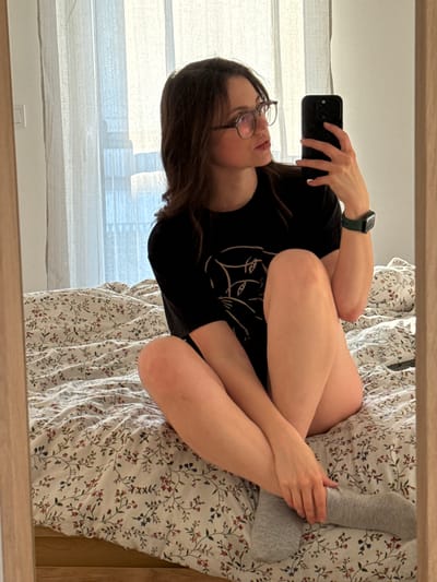 A person is reflected in a mirror. The person has brown hair and wears glasses, a black t-shirt with a graphic, and a smartwatch. They are holding a phone. The person is sitting on a bed covered with a floral patterned sheet and one leg is clad in a grey sock. Behind the bed is a window with white curtains. The location and city in Hungary are not apparent from the visual information.