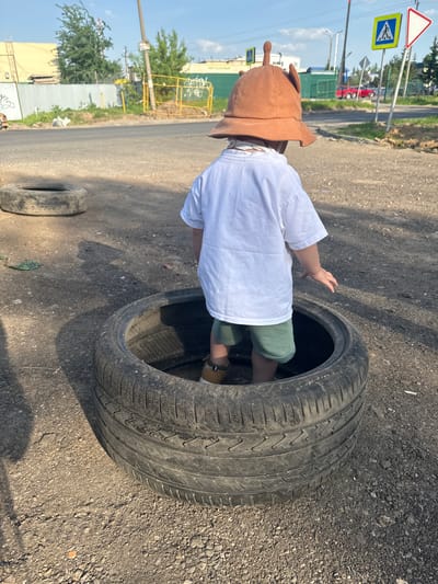 A child in a brown hat is standing inside a car tire on a gravel-covered surface in Akulovo, Gp Kubinka, Russia. The child is wearing a white shirt and green shorts. Another tire is lying on the ground nearby. A road with street signs is visible in the background. A yellow fence is adjacent to the road and behind it is a building. The sky is visible and clear.
