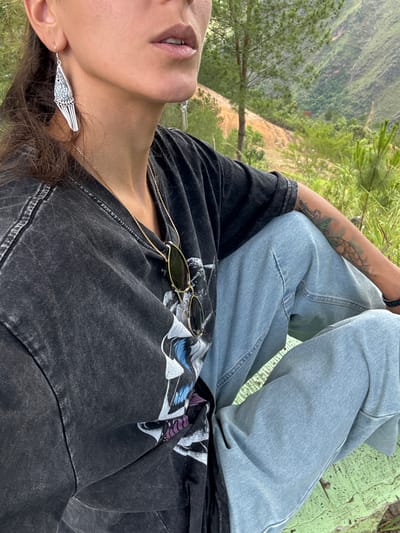 The image shows a person in Lũng Cú Commune, Vietnam, sitting outdoors with crossed legs. They are wearing light blue jeans and a black t-shirt with a graphic print, along with silver earrings and sunglasses. A gold chain necklace is visible. The background features green vegetation and a distant landscape. The person has a tattoo on their forearm. The image is rotated to the left.