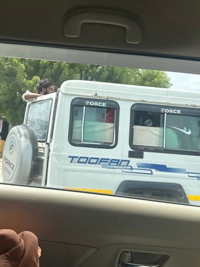 A man with dark, curly hair and a beard leans out of the open driver's side window of a white Force Motors vehicle. The vehicle has "TOOFAN" printed on its side. The man's left arm rests on the edge of the window frame. The spare tire is mounted on the rear left side of the vehicle, with "FORCE MOTORS" printed on its cover. A Nike swoosh logo is visible on the tinted rear window. The scene is set outdoors, with green trees visible in the background under a cloudy sky. The photo is taken from inside another vehicle, looking out through the windows. The interior of the observing vehicle is visible in the foreground, with a textured door panel and a portion of a person wearing a brown hooded garment.