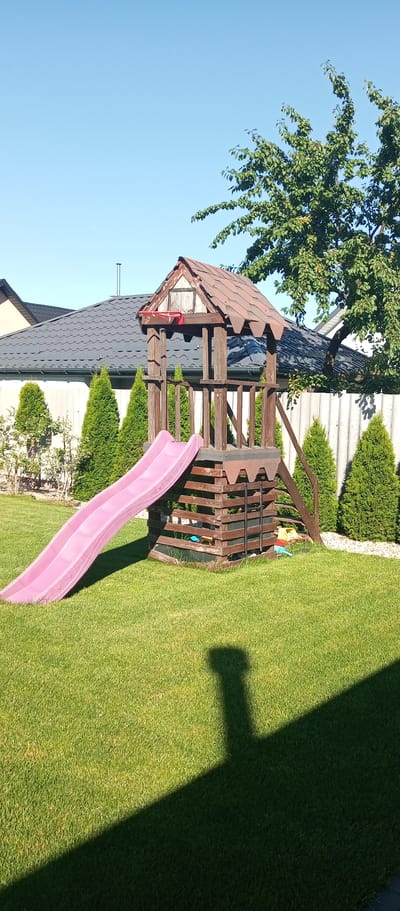 A wooden playground set with a pink slide is situated on a bright green lawn in a backyard. The playground is made of weathered wood and has a brown, shingled roof. A chain-link fence is visible in the background, behind which are several conifer trees. A portion of a building with a dark, wavy roof is also visible on the left side of the image. The sky is a clear, bright blue. A shadow of the playground structure is cast on the lawn. There are no people visible in the image. The scene appears to be in the city of Brest, Belarus.
