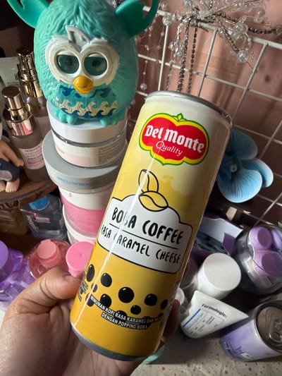 A hand holds a tall, yellow can with the "Del Monte Quality" logo and text that reads "Boba Coffee Rasa Caramel Cheese" and "Minuman Kopi Rasa Karamel Dan Ke... Dengan Popping Boba". The can is partially wrapped in clear plastic. In the background, various cosmetic products, including jars and bottles, are stacked on shelves. A turquoise toy with large eyes and wings is visible on top of some of the jars. Other items in the background include a blue fabric flower, decorative beads, and a silver grid. The setting appears to be a shelf or display within a home or shop, likely in East Jakarta, Indonesia, given the text on the can which appears to be in Indonesian.