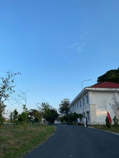In Lâm Viên - Đà Lạt, Vietnam, a clear blue sky with scattered wispy clouds is visible above a landscape featuring a long, white, multi-story building with a red tiled roof. The building has numerous windows and appears to be an educational or institutional facility. A paved asphalt road curves into the foreground from the right, leading towards the building. On the left side of the road, there is a grassy area with scattered young trees and dry branches. Three individuals, appearing to be young men dressed in light-colored clothing, are standing on the road near the building, engaged in conversation. A bright red flag is affixed to a pole on the side of the building. The environment suggests a temperate climate, possibly late afternoon due to the lighting.
