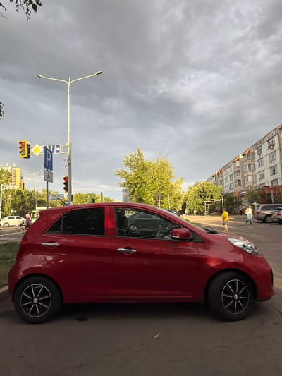 A red hatchback car is parked on the side of a street in Astana, Kazakhstan, under a cloudy sky. In the background, a multi-story residential building with a distinct architectural style lines the street. Across the road, several pedestrians are visible, including a person in a yellow t-shirt and shorts walking across the street, and a woman with a bag. Trees with green foliage line the sidewalks and are interspersed with streetlights and traffic signals. A white car is seen in the distance on the left side of the frame, near a traffic light. The overall scene depicts a typical urban environment in Astana during daytime.