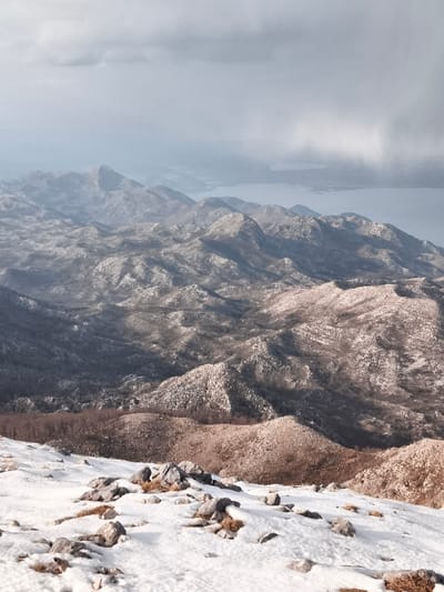 The image displays a panoramic, high-angle view of a snow-dusted mountainous landscape, identified as Mali Mikulići, Montenegro, under an overcast sky. The immediate foreground shows patchy white snow covering uneven ground, interspersed with exposed gray rocks and sparse, dry brown vegetation. Extending into the midground, numerous undulating hills and mountains dominate the scene, exhibiting a mixed cover of snow, bare deciduous trees, and exposed dark earth and gray rock. Shadows define the various contours and slopes of these formations. In the distant background, a calm body of water, likely a lake or bay, is visible, bordered by additional mountain ranges that gradually fade into a light blue-gray atmospheric haze. The sky overhead is cloudy, ranging from light to dark gray, with a subtle brighter area in the upper right quadrant. No individuals, man-made structures, or active interactions are present in the frame.