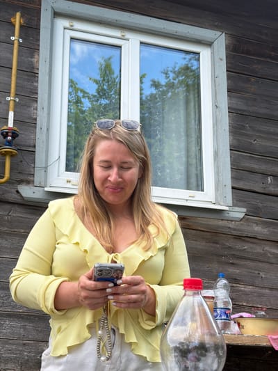 A woman with blonde hair and sunglasses perched on her head, wearing a yellow ruffled blouse and white pants, stands in front of a wooden building with a window. She is looking down at her smartphone, which she holds in her hands. Beside her on a table are a bottle of water, a large plastic bottle with a red cap, and a bowl. To the left of the woman, a yellow pipe with a valve is attached to the building. The background shows trees and the sky reflected in the window. The scene appears to be outdoors in a rural or suburban setting, possibly in Бронница, Russia.